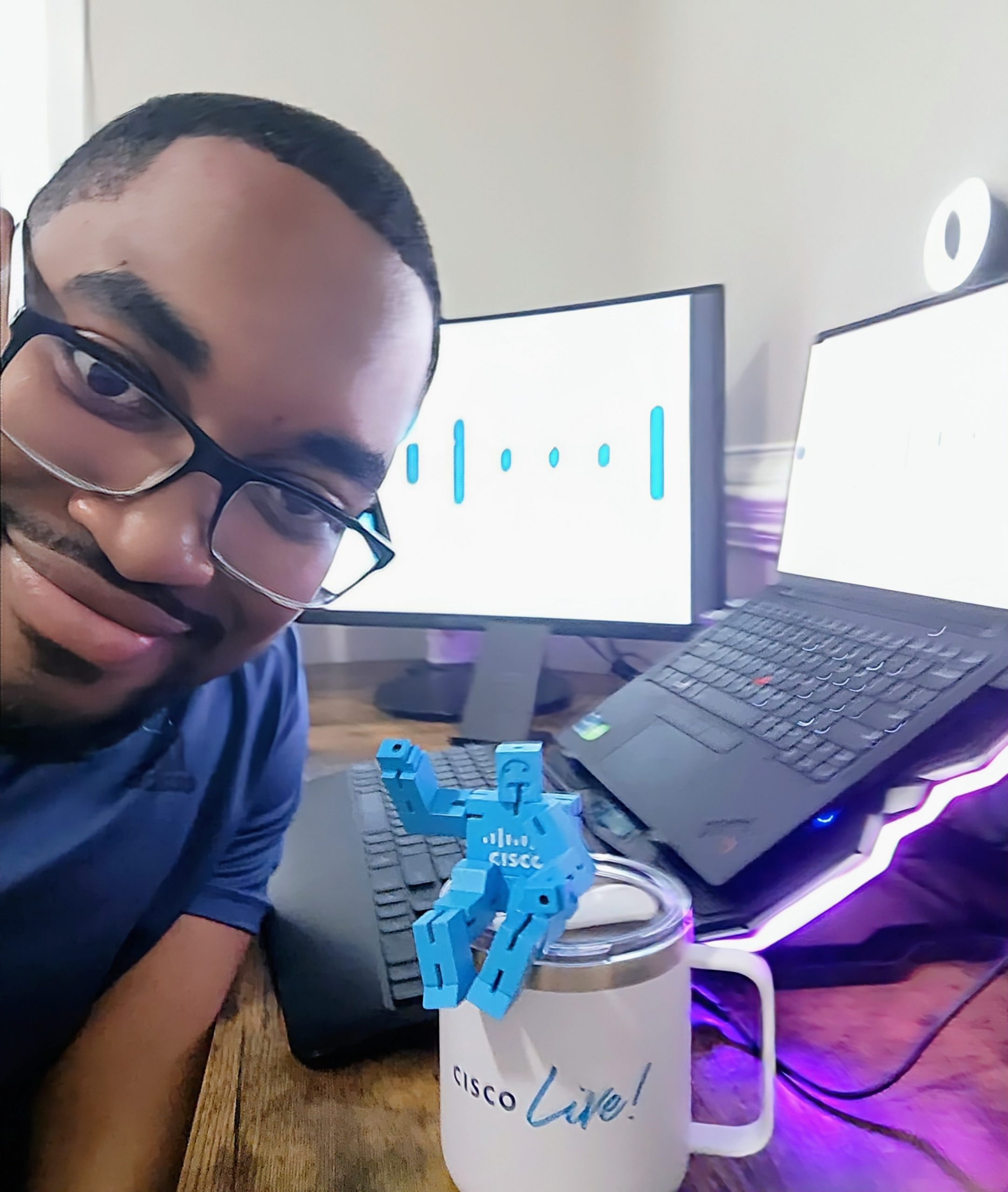 Dark-skinned man with dark hair and black-rimmed glasses taking a selfie at his desk with Cisco Live white coffee mug and blue Cisco lego man with monitors in the background displaying the Cisco logo.