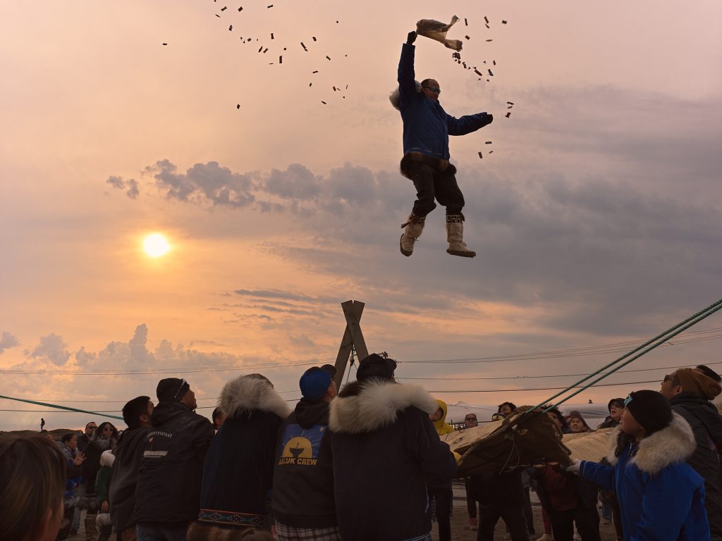 Residents of Utqiaġvik take part in the traditional blanket toss with person high in the air with candy flying our of bag.