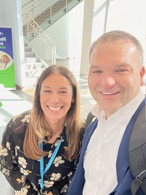 Light-skinned man and woman, both with brown hair, take a selfie in office building with white staircase behind them.