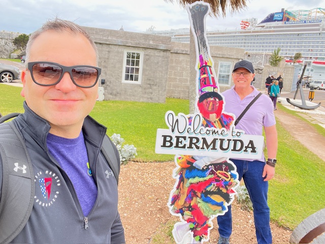 Two men taking a selfie by a "Welcome to Bermuda" sign with a gray stone building and cruise ship in the background.