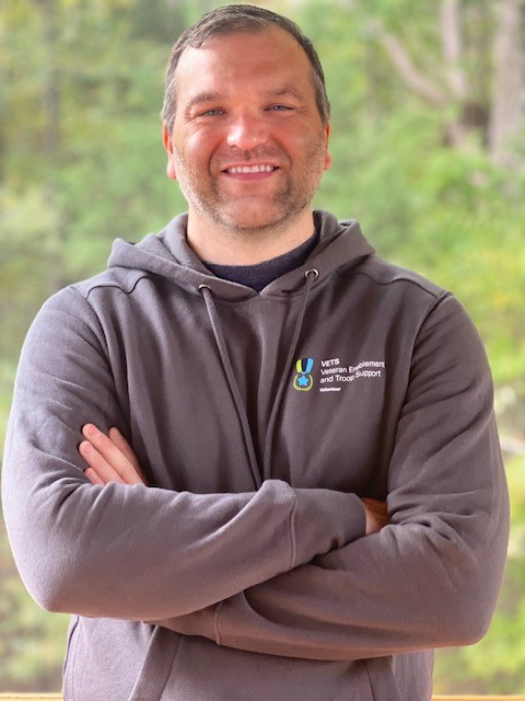 Light-skinned man with brown hair, posing with arms crossed, wearing gray Cisco VETS Inclusive Community hoodie with blurred trees behind him. 