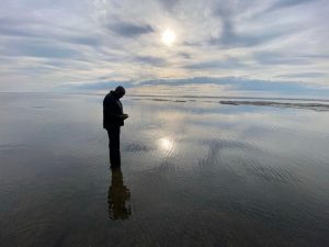 Scott McGregor standing at water's edge against cloudy sky with sun shining.