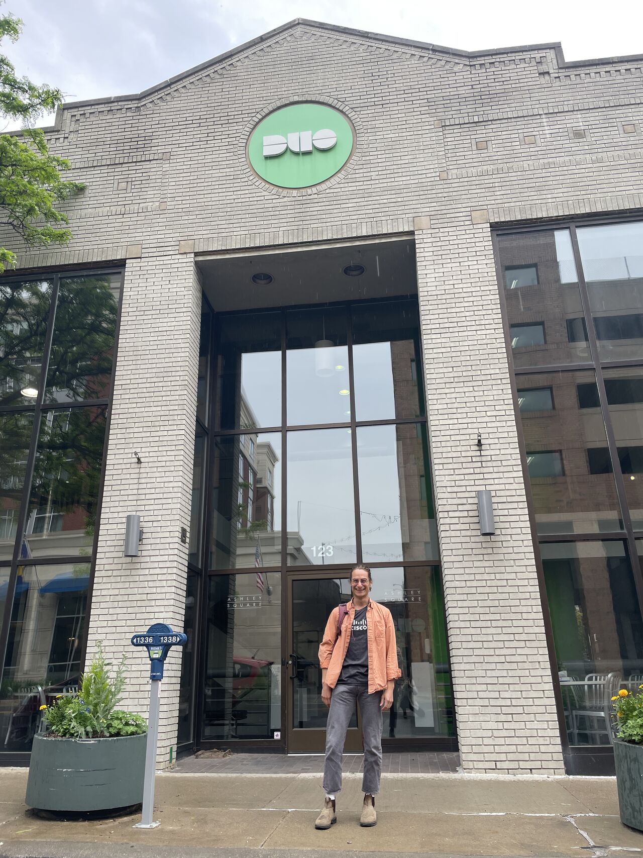 Light skinned young man wearing gray pants, Cisco t-shirt, orange button up shirt, and backpack posing in front of building with green DUO logo.