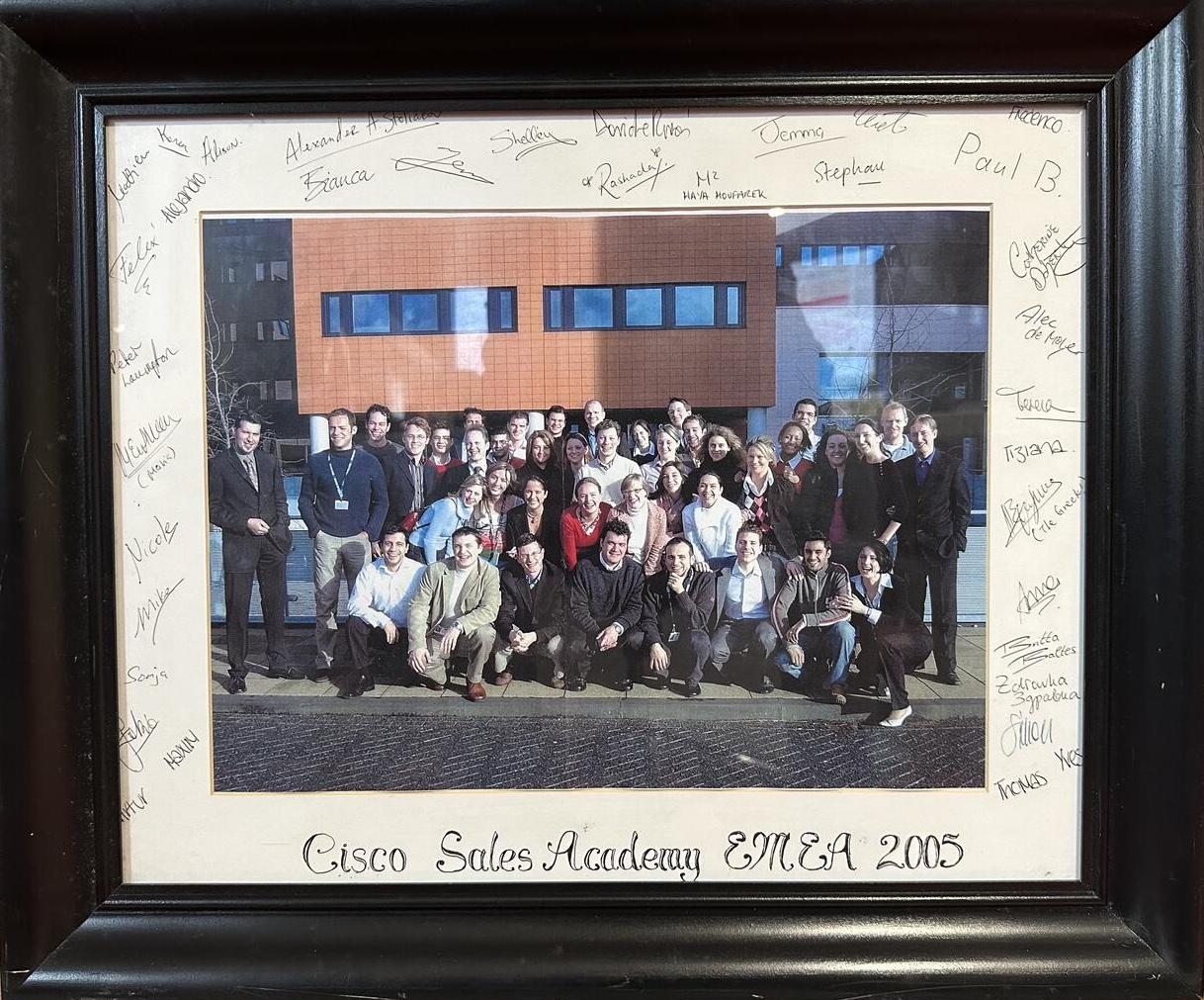 Framed group photo where they are posing in front of an office building. It's in a black wood frame with cream mat and there are signatures on the mat and under the photo text reads "Cisco Sales Academy EMEA 2005."