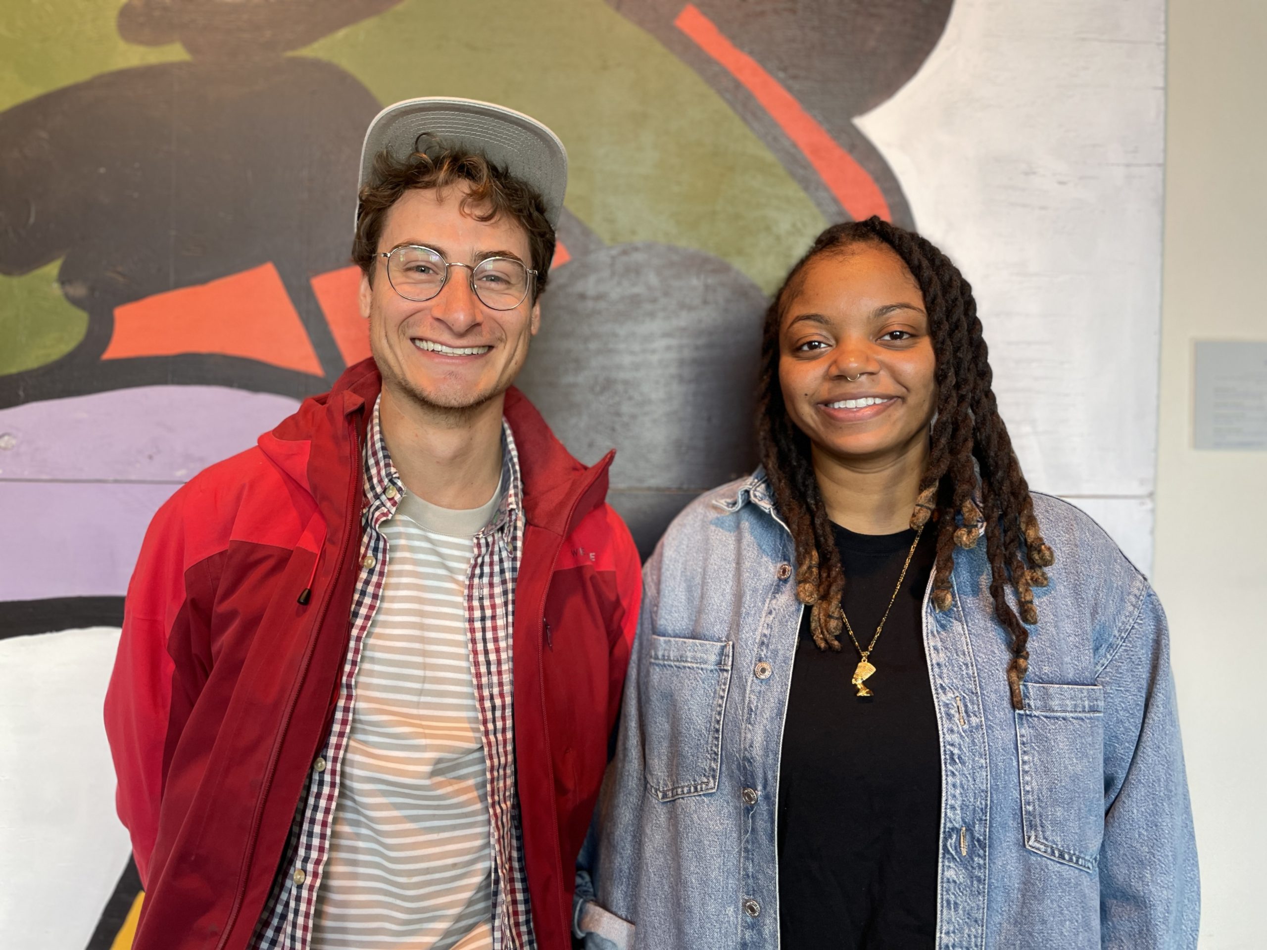 A light skinned young man wearing red jacket and white hat and dark skinned young woman wearing denim shirt pose smiling against mural.