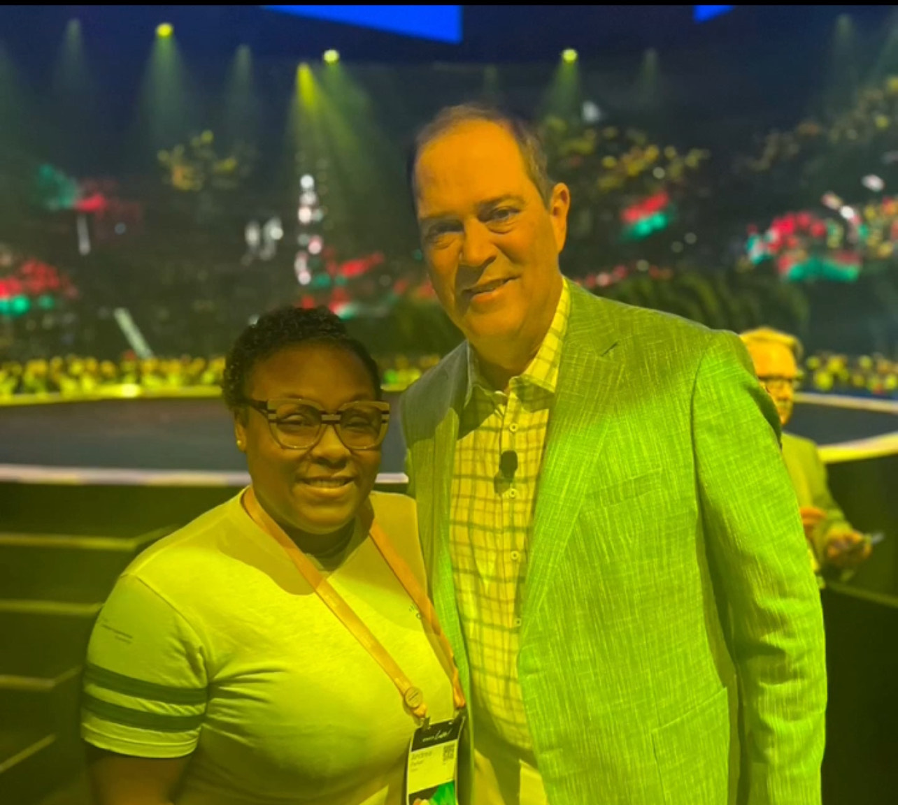 Short, dark-skinned woman with short dark hair and glasses wearing t-shirt posing with taller light-skinned man with dark hair, button-up shirt and sport coat, yellow-green light shining over both of them.
