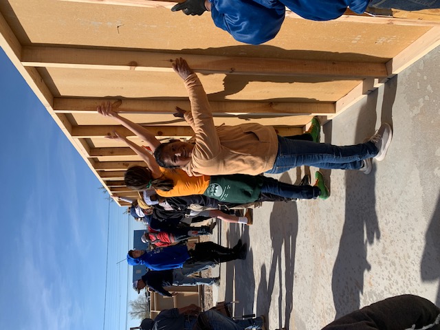 Group of people holding up framed wall for house at building site.