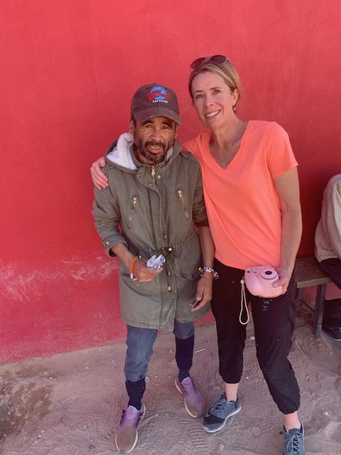 Man wearing green jacket, tennis shoes, and baseball cap, and woman wearing black joggers and neon orange shirt standing, smiling against red wall.