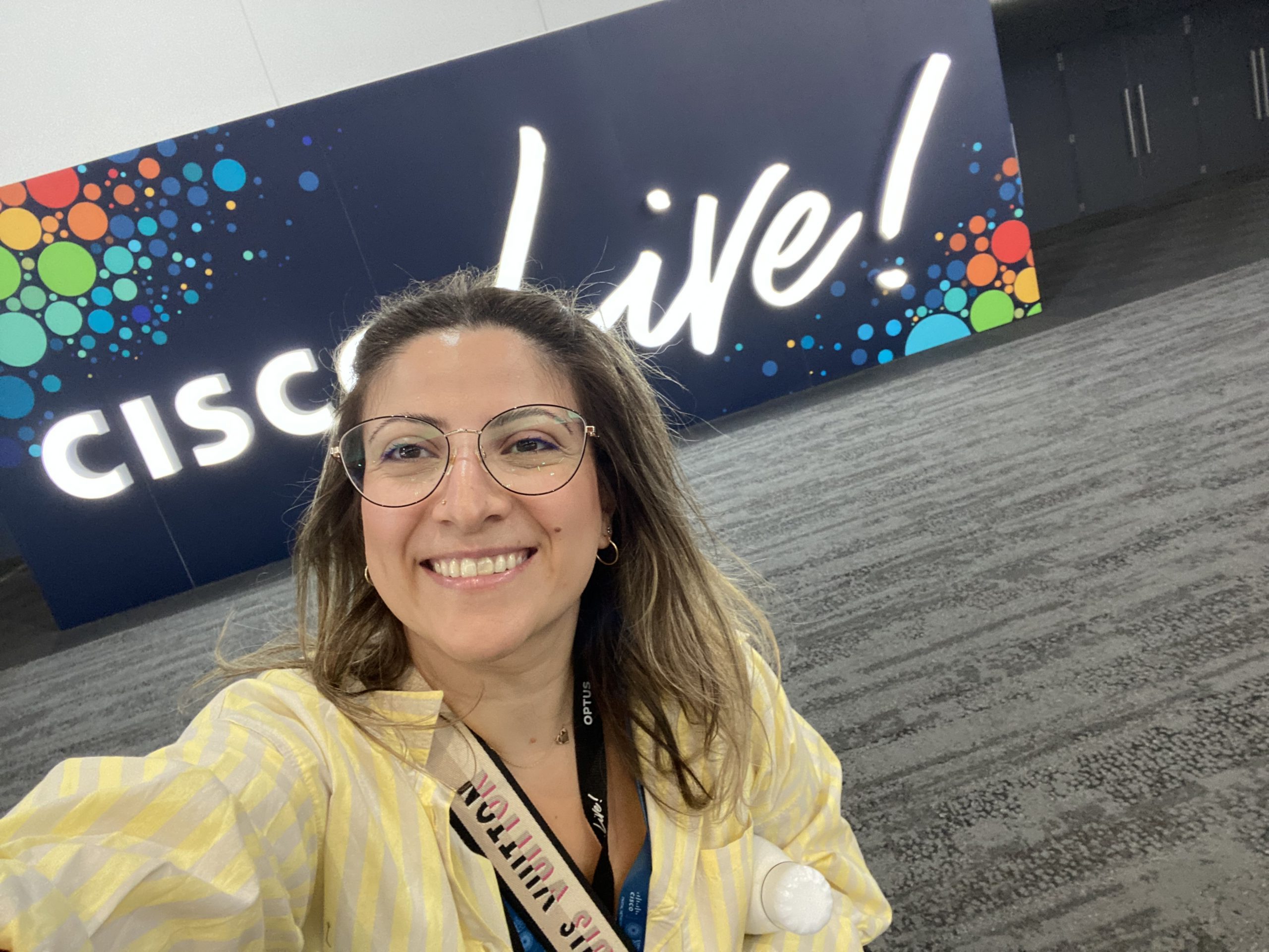 Woman smiling taking selfie wearing yellow blouse and glasses with Cisco Live sign in the background. 