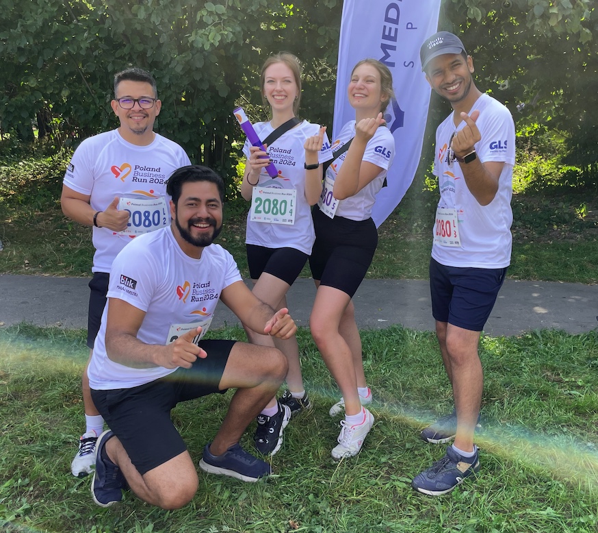 A group of five people in athletic wear, posing cheerfully outdoors after a race, wearing numbered bibs and smiling with thumbs up.