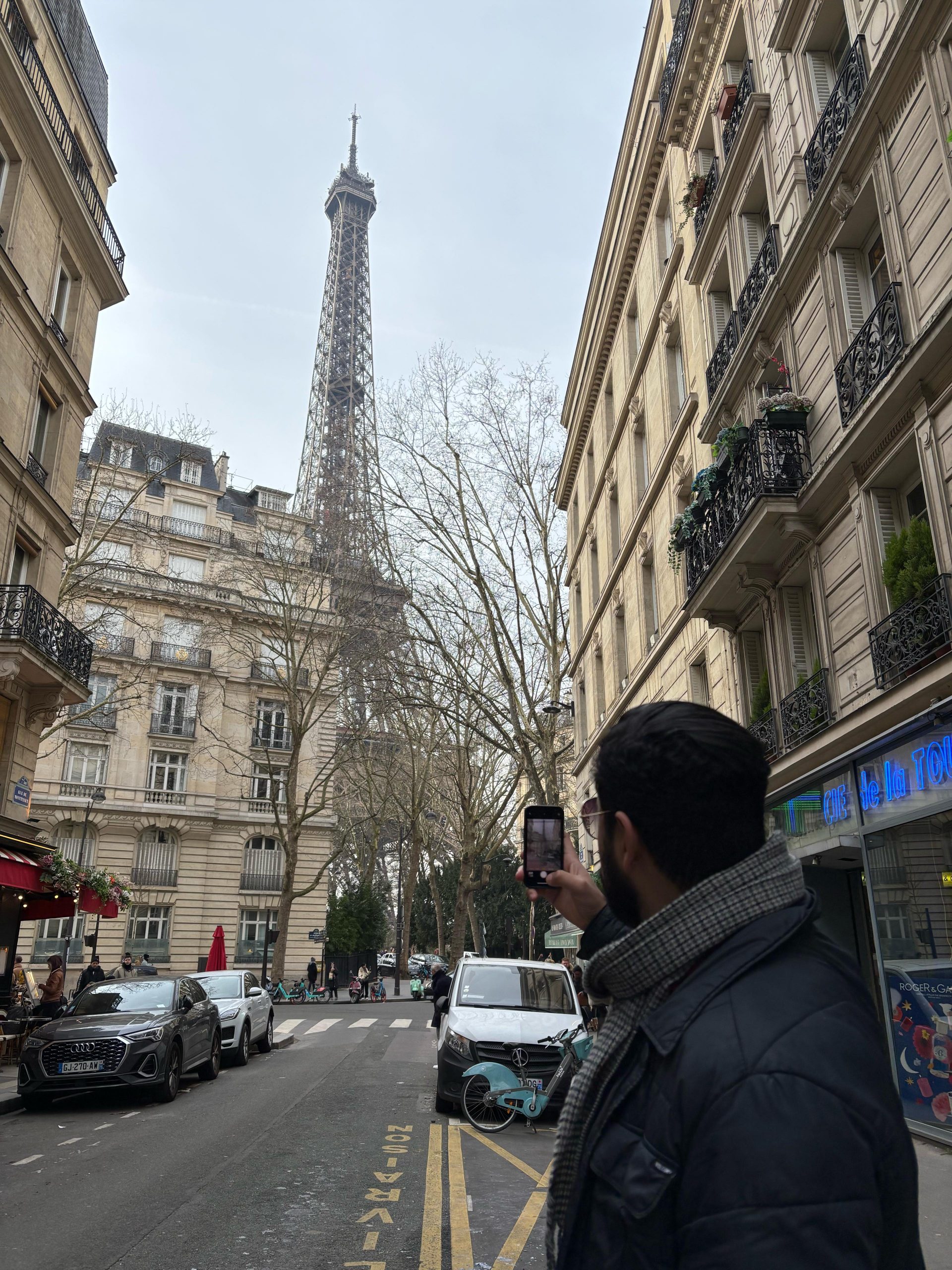 A person stands on a street in Paris, taking a photo of the Eiffel Tower on a cloudy day, surrounded by classic Parisian buildings.