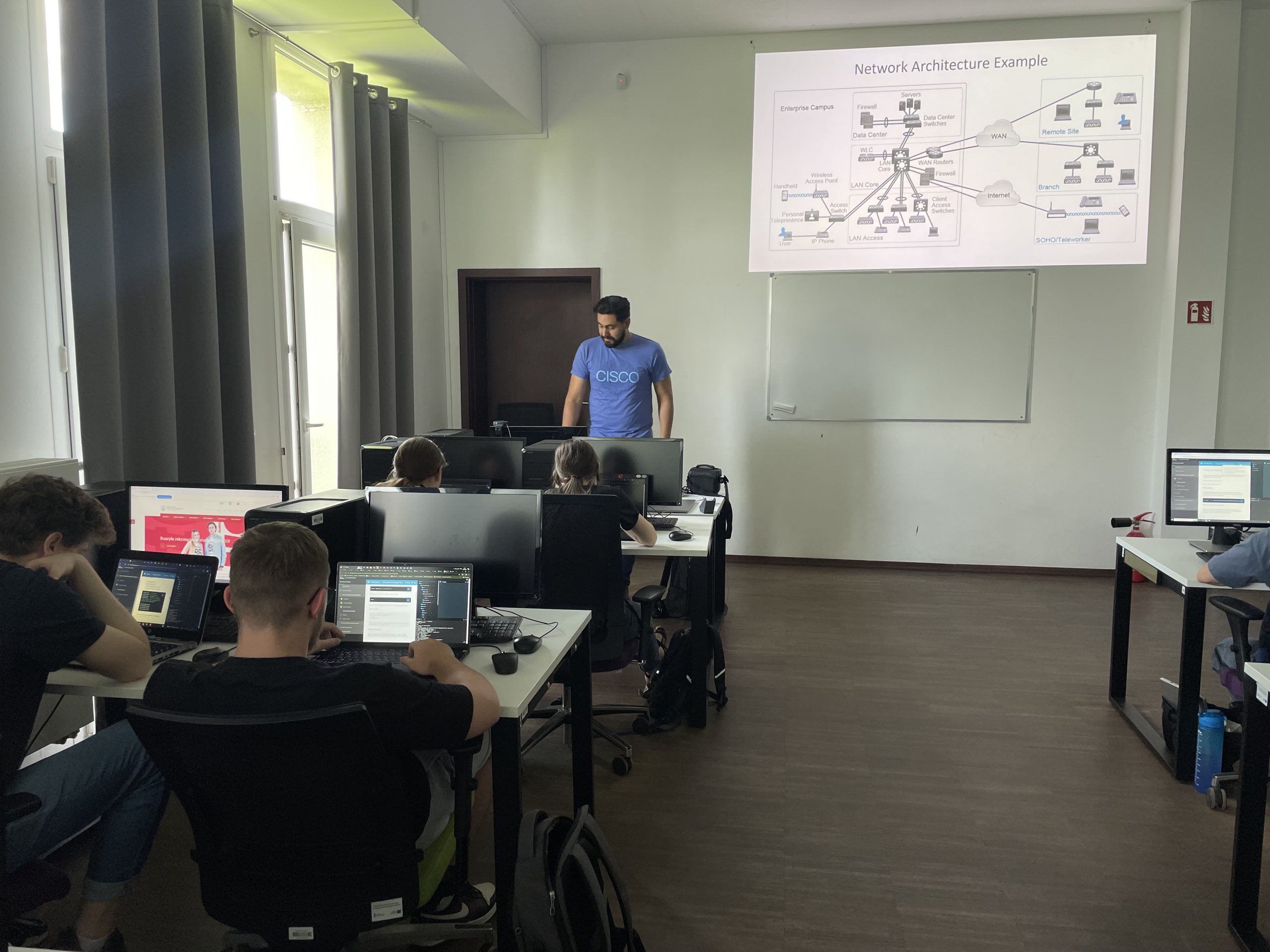 Students in a classroom use laptops while an instructor in a blue shirt stands near a whiteboard with a diagram, in a room with large windows, a wooden floor, a blue checkmark in the top right, and a cloud icon in the bottom right.