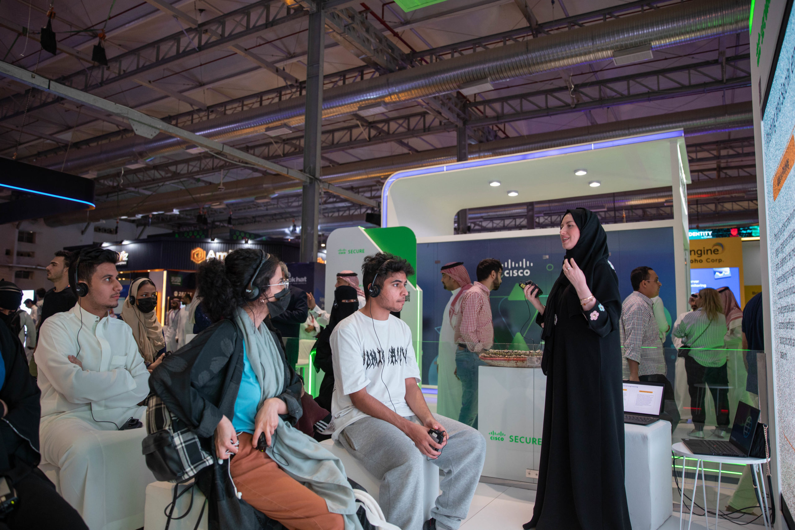 A woman in a black headscarf presents to a seated group of young attendees at a tech event. The audience listens attentively as she speaks in front of a modern exhibition booth with bright lighting and digital displays.