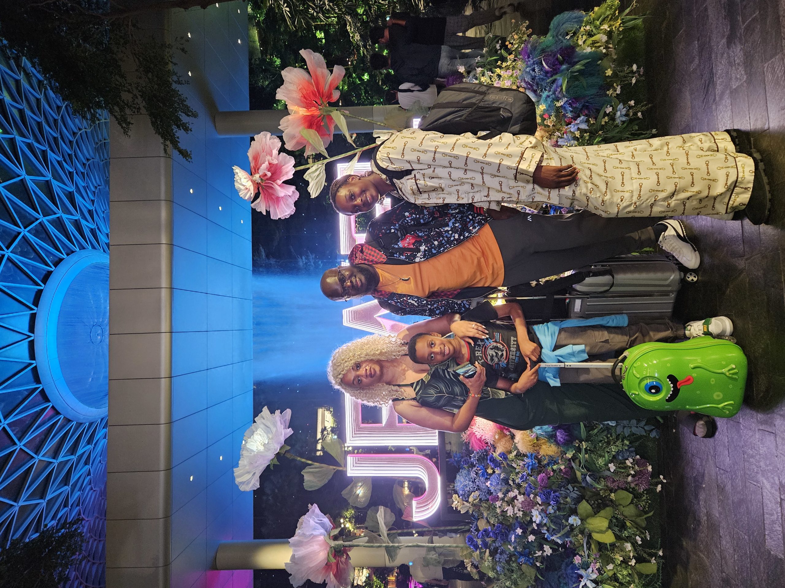 A family of four, including two adults and two children, stands in front of a large illuminated sign and colorful floral decorations. They are standing next to several suitcases, including a bright green monster-shaped suitcase.