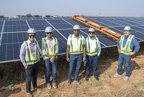 Five people wearing hard hats and yellow vests standing in front of solar panels.