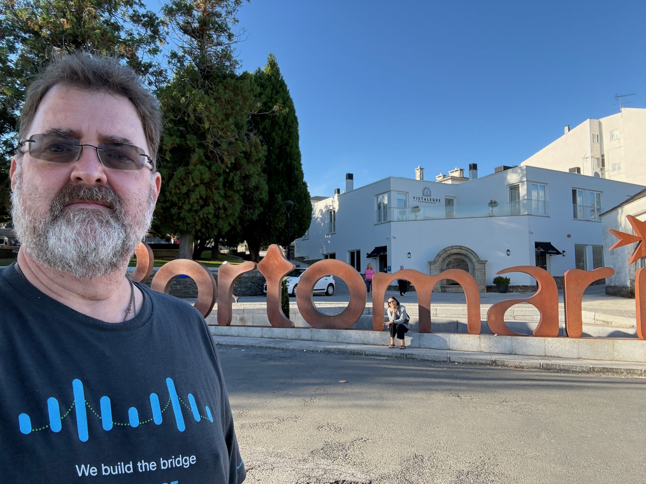 Light-skinned man with brown hair and salt and pepper beard and glasses, wearing a black Cisco "We build the bridge" t-shirt takes a selfie in front of large free-standing Portomarín sign in Spain with white building in background.