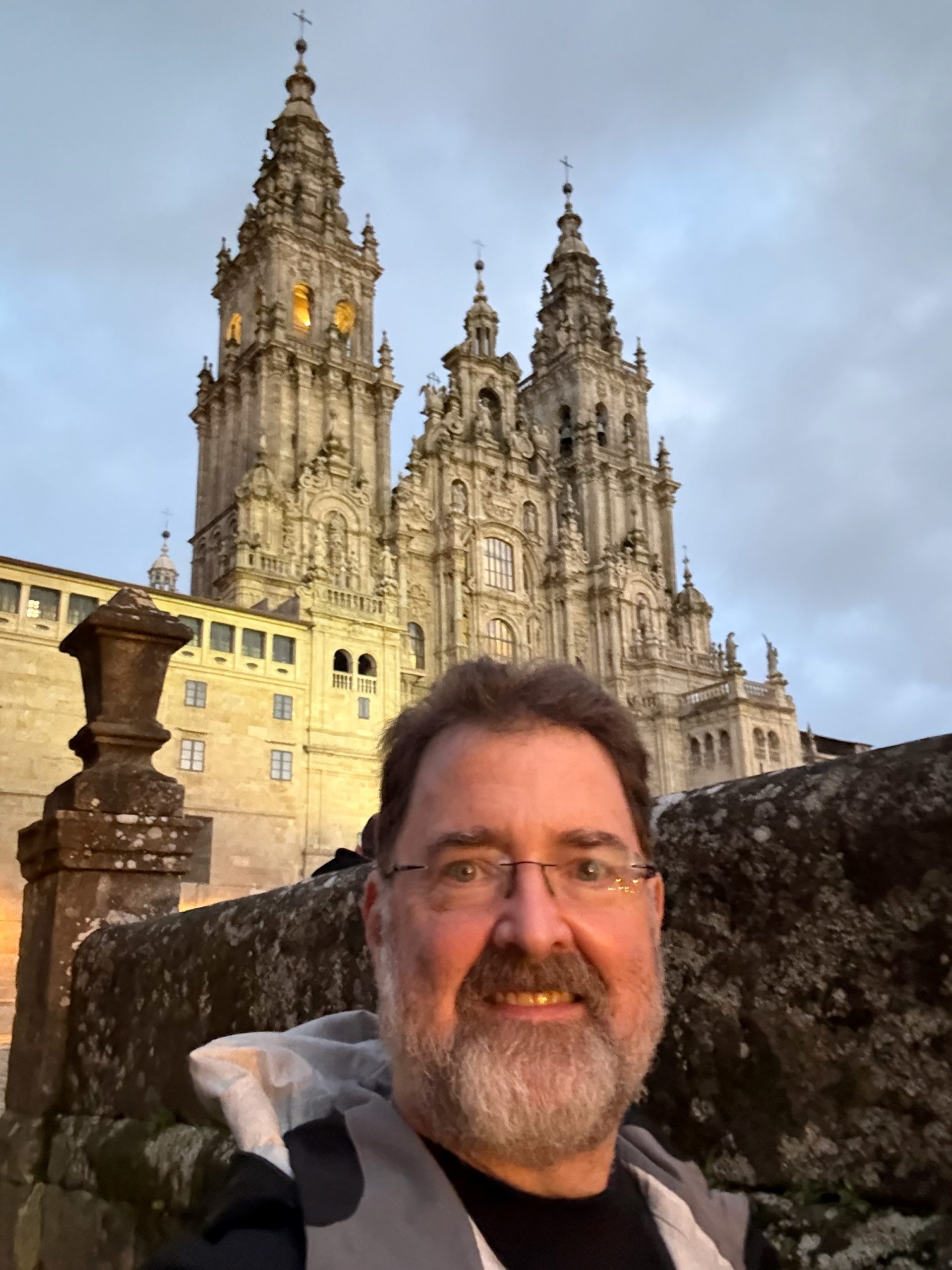 Light-skinned man with brown hair, salt and pepper beard, wearing glasses and a jacket takes a selfie in front of Santiago de Compostela Cathedral in Spain.