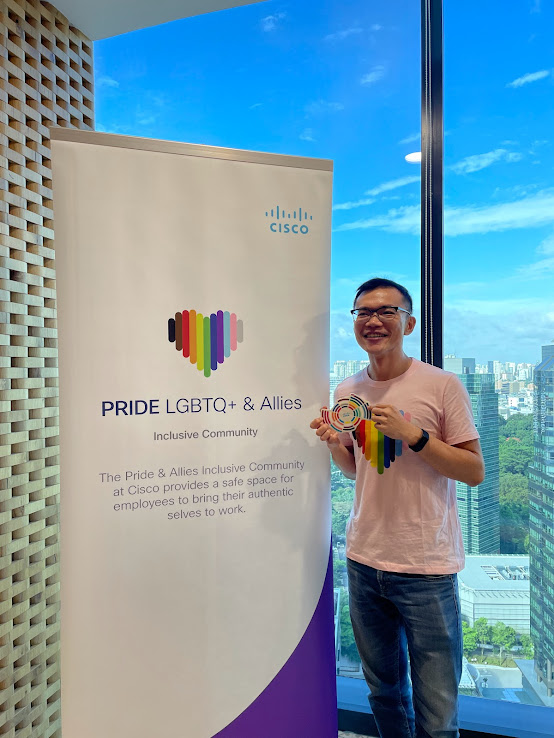  A person standing in front of a "Pride in Asia" banner with a rainbow heart logo, indoors with a cityscape visible through the window. 
