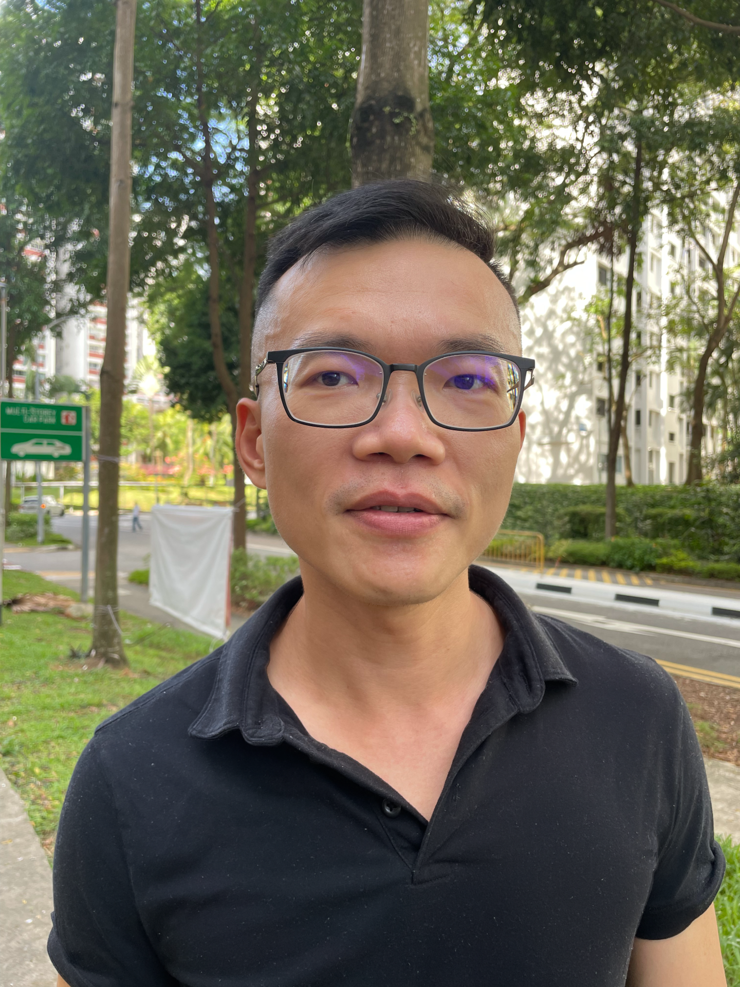 An eye-level, outdoor portrait shows a man with short dark hair and black-framed glasses looking directly at the viewer. He has light skin and is wearing a black polo shirt. The background is out of focus but shows green trees, a paved road, a green street sign, and white buildings.