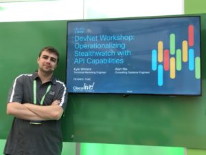 A tall man with light skin wearing a Cisco polo shirt stands next to a TV screen that contains text about DevNet