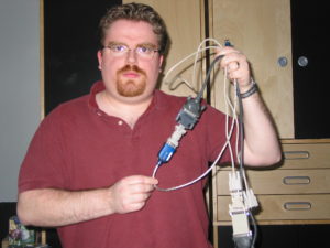 A light-skinned man with a brown goatee wearing a red polo shirt holds a bunch of cords