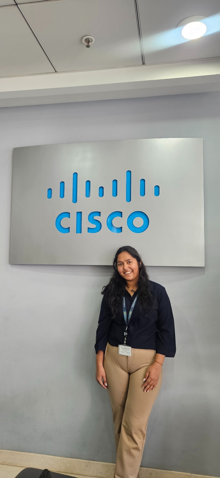 Woman with Black hair, black button down shirt and khaki pants standing in front of a big Cisco sign.
