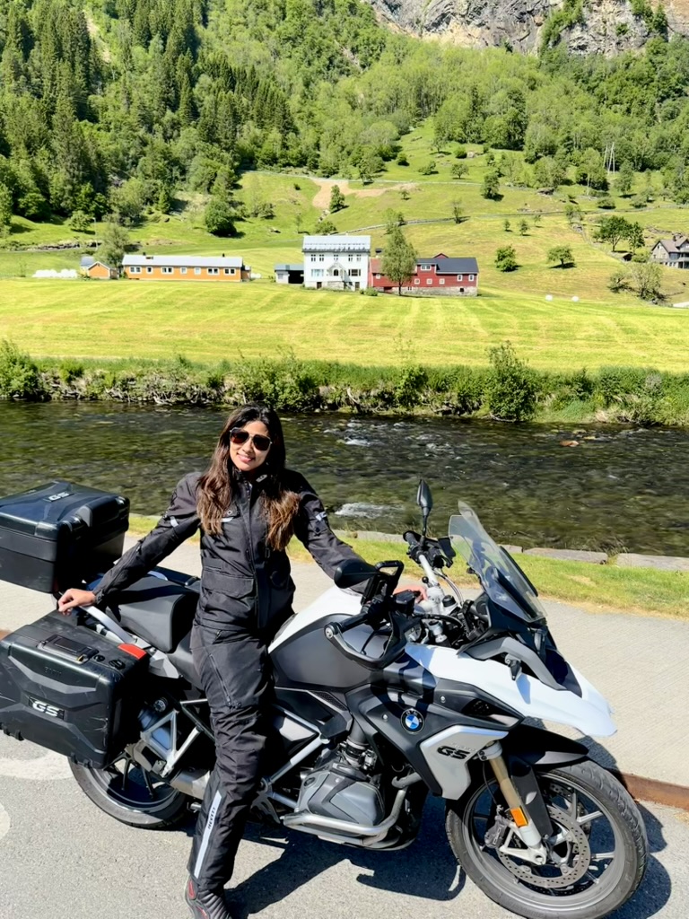 A woman with long, dark hair in black motorcycle gear sits on a black motorcycle, with a river, green fields, houses, and forested mountains in the background.