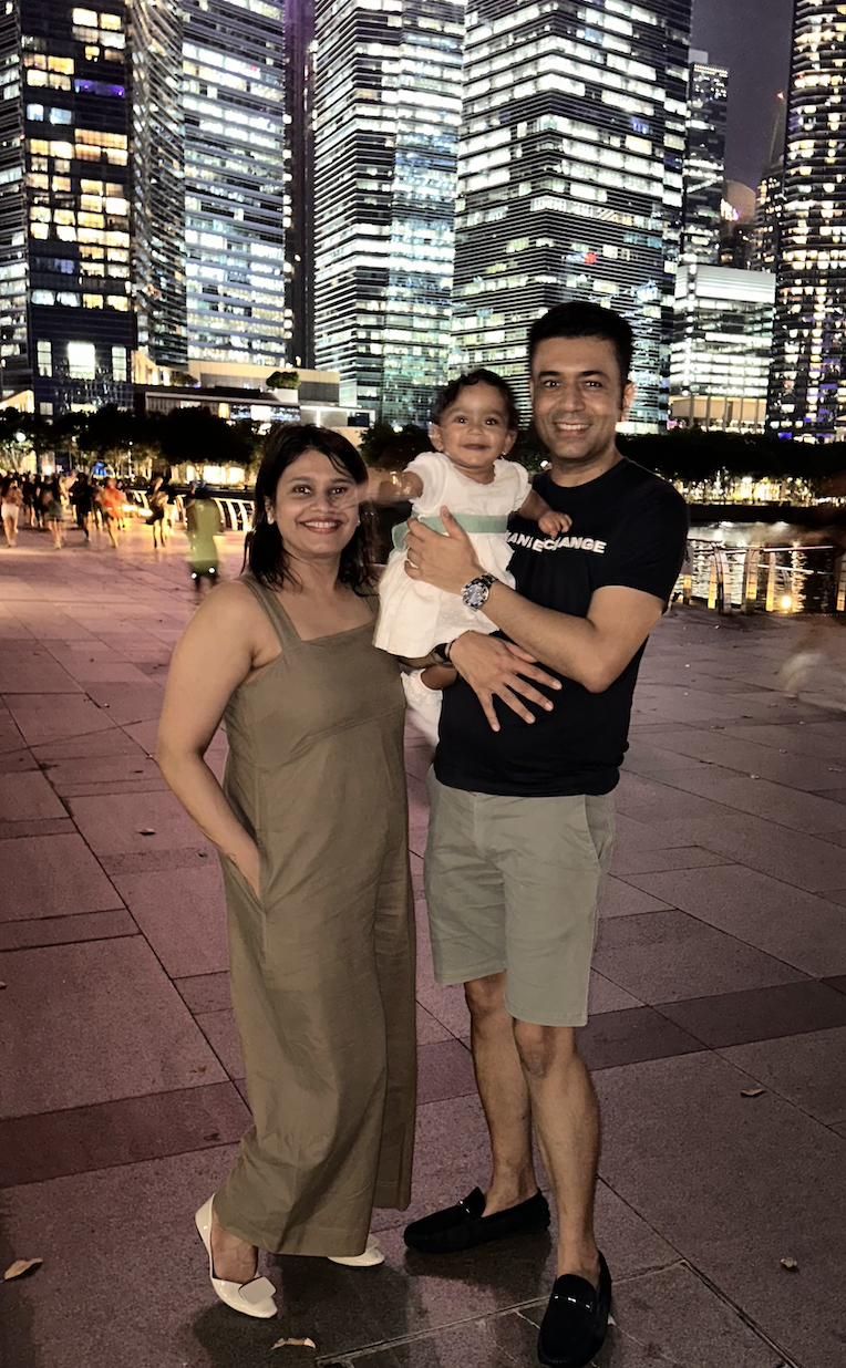 A smiling family of three (man, woman, and baby) standing outdoors at night, with illuminated city skyscrapers in the background.
