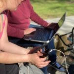 Two women on a park bench on a computer and a laptop.