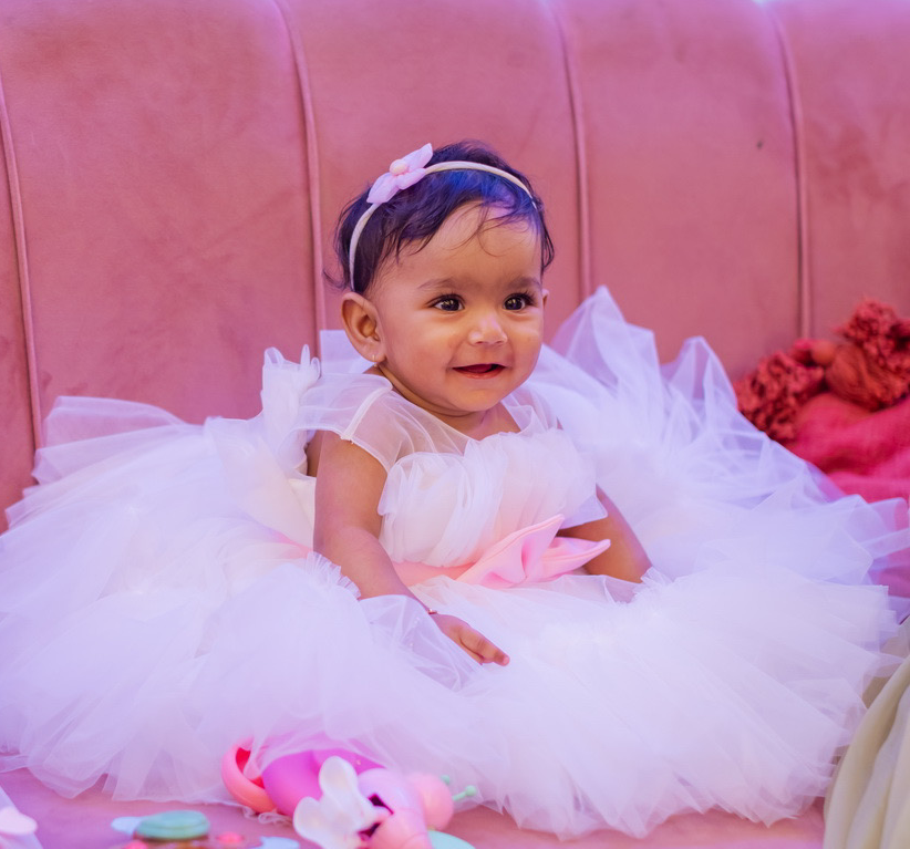 A baby in a white and pink dress, wearing a headband with a flower, smiles while sitting on a pink couch.