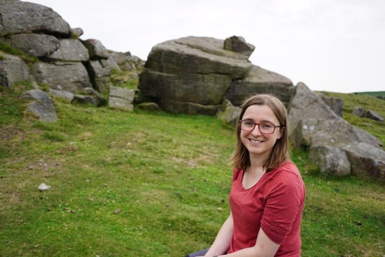 A smiling woman in glasses and a red shirt sits in a grassy, rocky landscape.