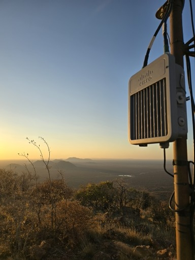 A Cisco outdoor networking device stands prominently on a hilltop, overlooking a vast, scenic landscape bathed in the warm, golden light of either sunrise or sunset.