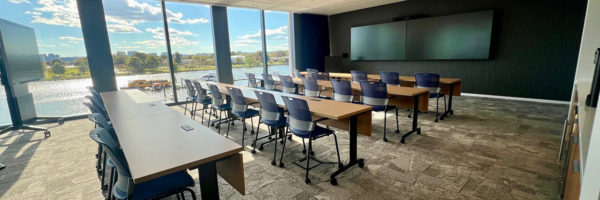 A spacious, modern multi-purpose room at Cisco Georgetown, with tables and chairs arranged for a meeting, offering a panoramic view of the water and cityscape through large windows.