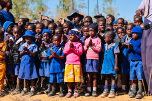 A large group of young children in blue uniforms stand outdoors, many with hands pressed together.