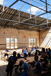 Children are seated at desks and gathered in a rustic classroom with open windows and an exposed roof structure.
