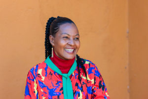 A smiling woman with braided hair wears a vibrant multi-colored jacket over a red turtleneck, looking to the side.