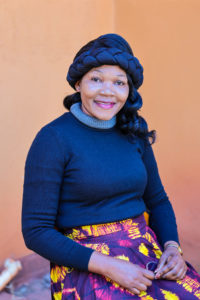 A woman with a braided headwrap and a colorful skirt smiles, wearing a dark blue sweater, against an orange background.