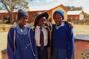 Three smiling young girls in school uniforms stand together outdoors in front of a school building.