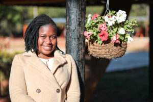 A smiling woman with braided hair wears a light brown coat, leaning against a wooden post with a hanging flower basket.