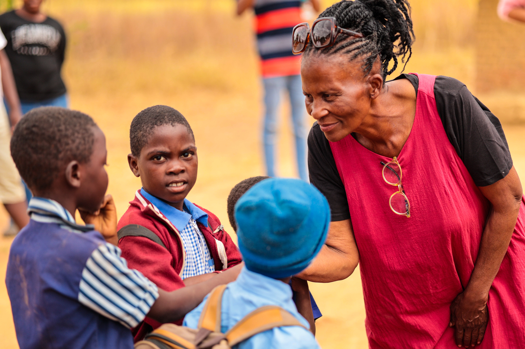 An adult woman in a red dress with sunglasses on her head smiles at three young children outdoors, one of whom looks directly at the camera.