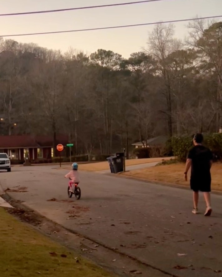 Man walking at dusk behind his daughter on a bicycle.