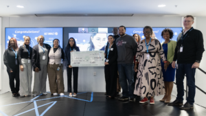 A group of people stand on a stage. Two women in the center hold a big check for the $10,000 first place prize.