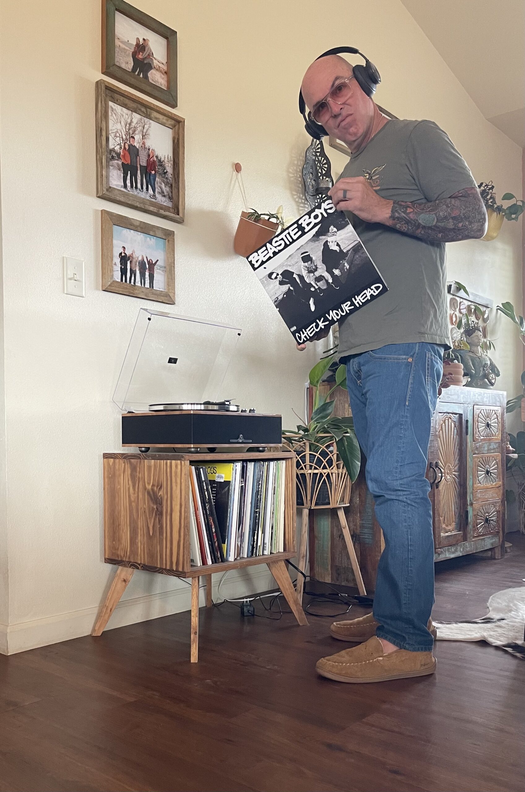 A man wearing headphones holds a Beastie Boys "Check Your Head" vinyl record next to a record player and a collection of albums in a living room.