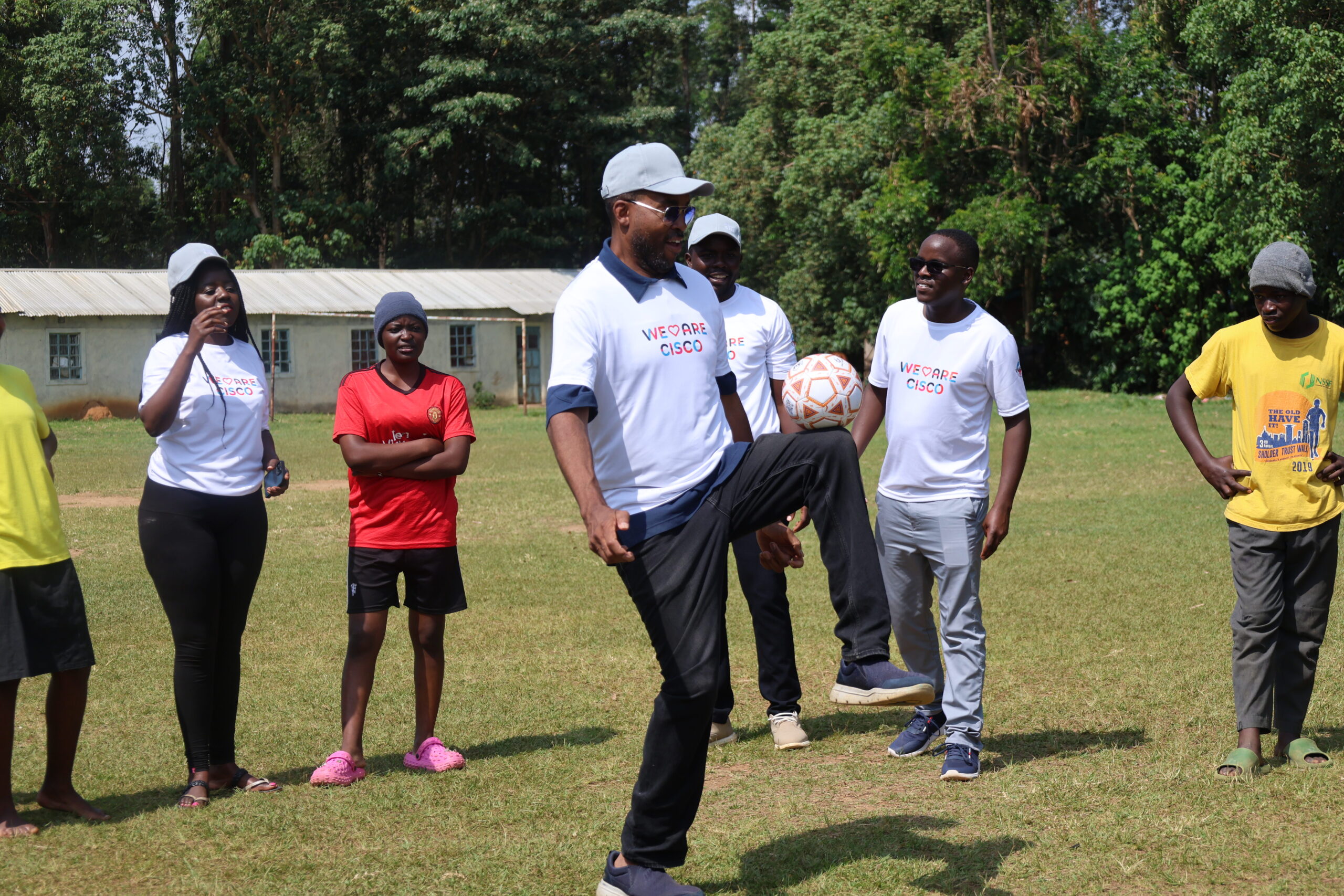 Man wearing a ‘We Are Cisco’ T-shirt balances a soccer ball on his knee while a small group watches on a grassy field.