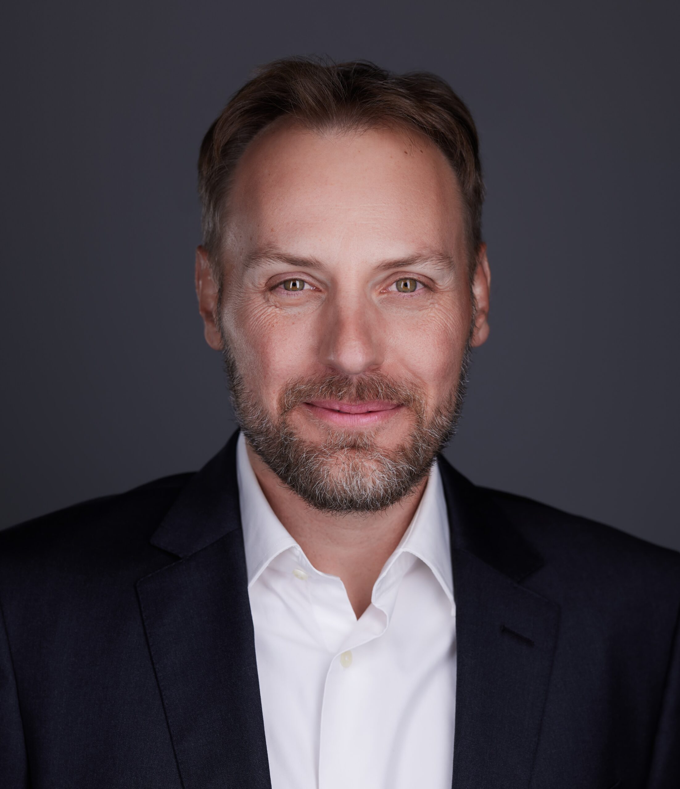 A man with a beard wearing a white collared shirt and black blazer