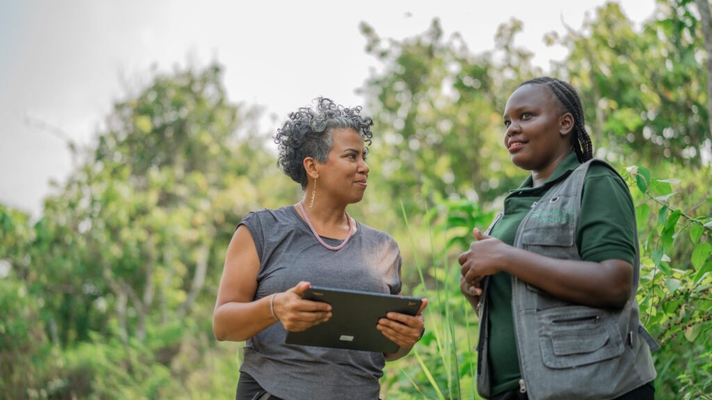 Image of 2 women speaking together in a field of green, holding a laptop. 