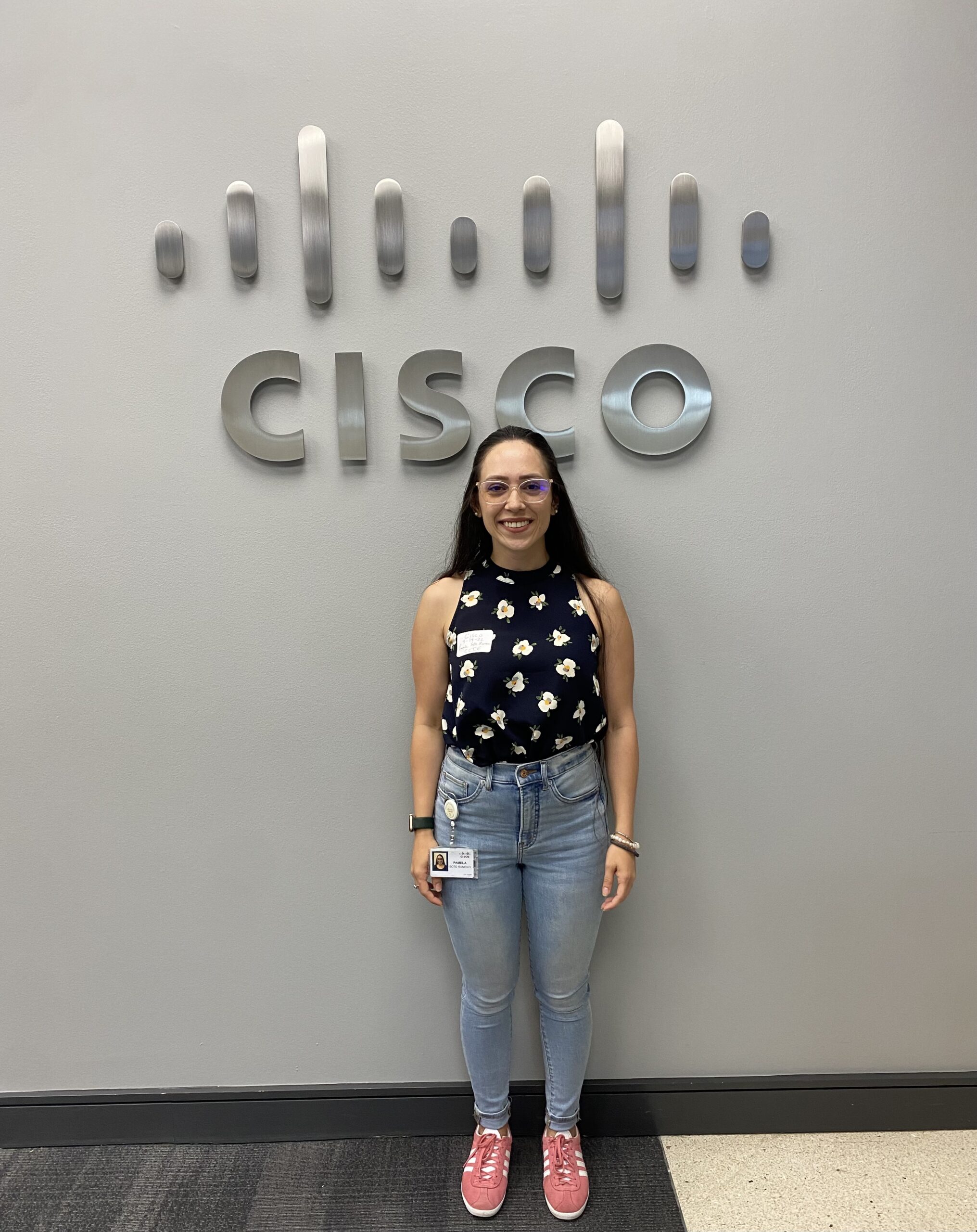 A woman stands centered in front of a gray wall with a metallic Cisco logo, wearing a floral sleeveless top, jeans, and pink sneakers, posing for a casual office portrait.