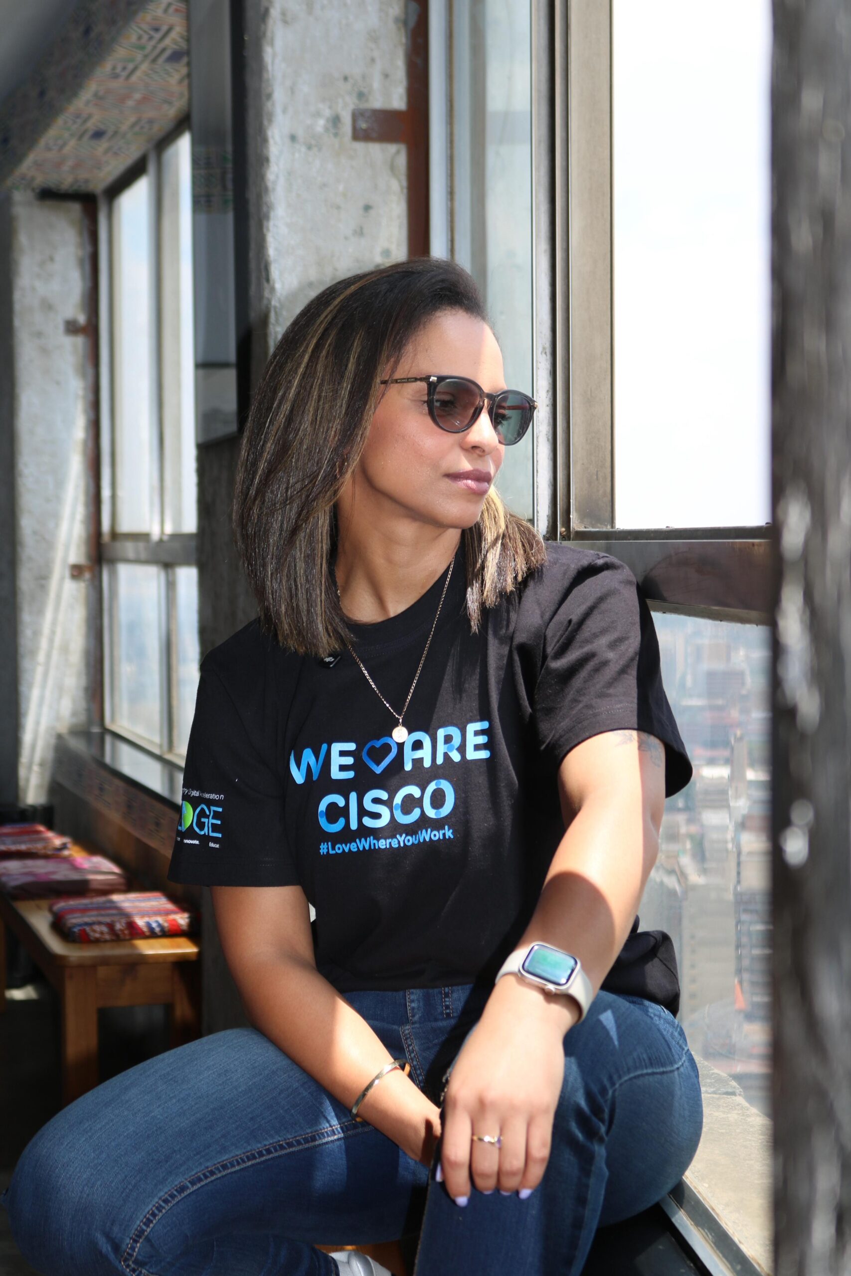 Woman wearing a “We Are Cisco” T-shirt and sunglasses sitting by a window with natural light.