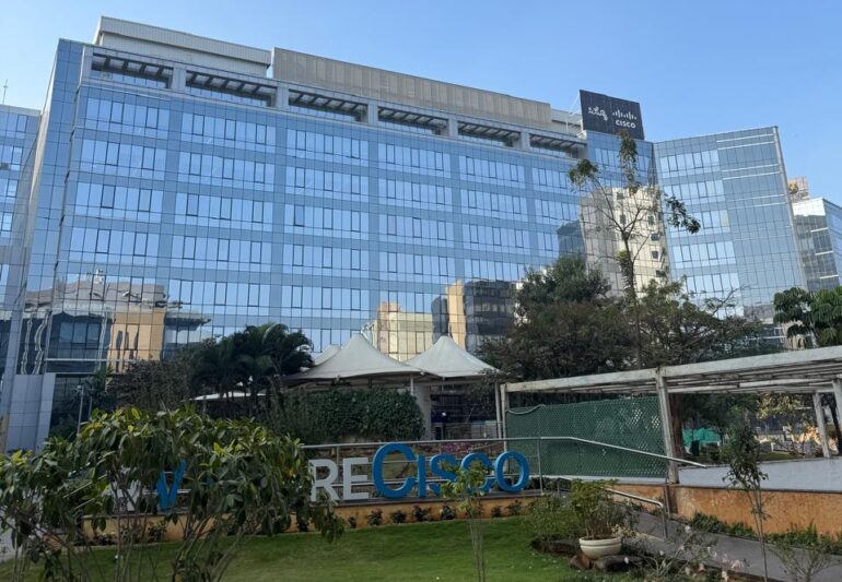 Daytime exterior of Cisco Bangalore - a tall, glass building with green lawn and #WeAreCisco sign in front