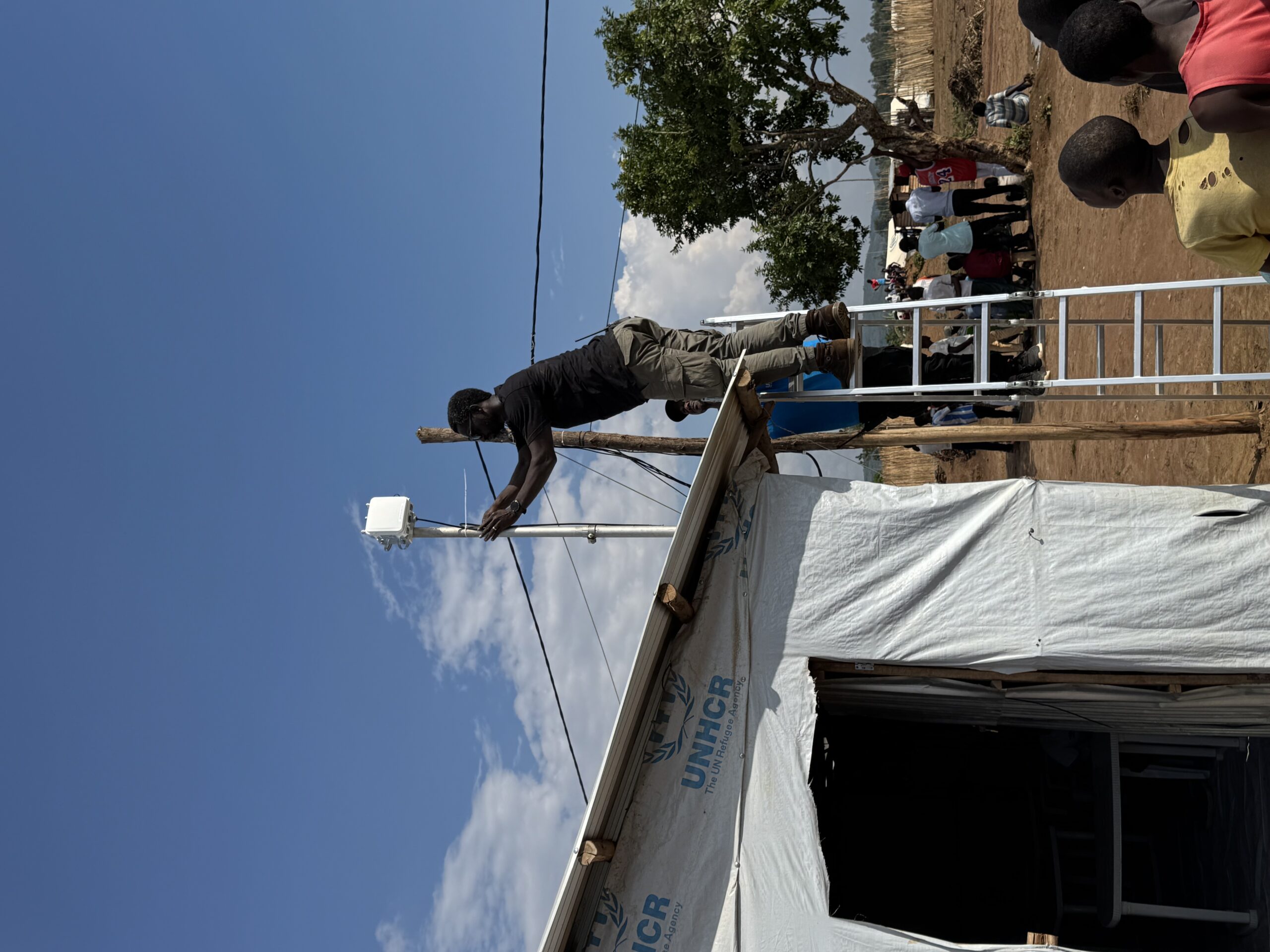 A man stands on top of a ladder to install an antenna on top of a UNHCR tent at a refugee site in Burundi. Several people look up at him to watch off to the right, while others sit under a tree in the background.